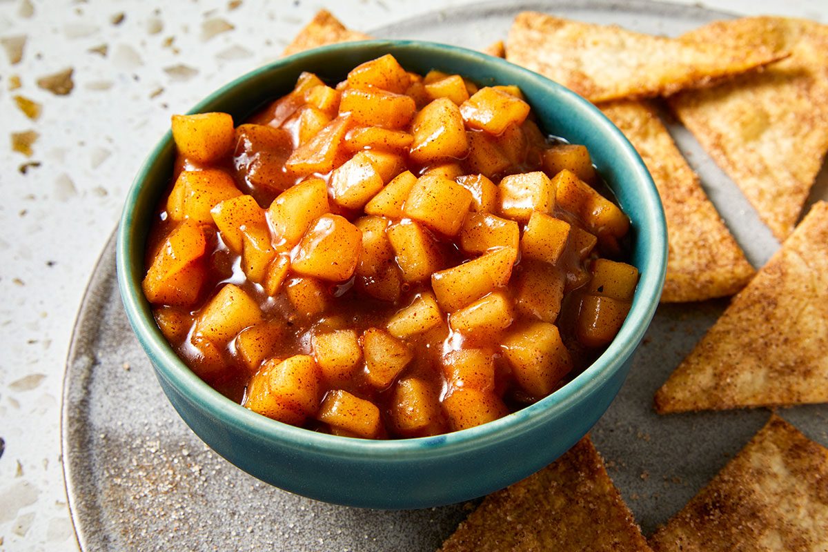 Closeup shot of a turquoise bowl filled with chunky apple pie dip coated in a glossy, spiced brown sauce, set on a plate with cinnamon-sugar pita crisps in the background;