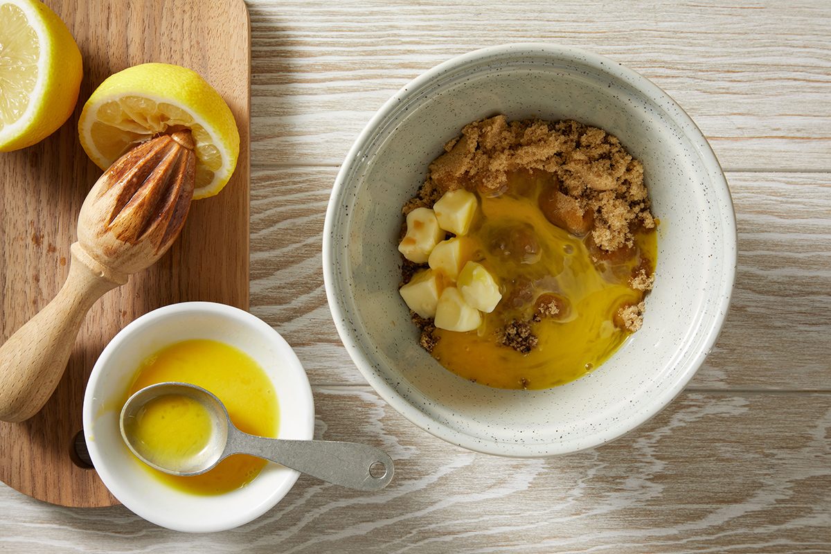 A bowl containing brown sugar, chopped banana, and cracked egg sits on a light wooden surface. Nearby are a halved lemon with a juicer on a cutting board and a small bowl of melted butter with a spoon.