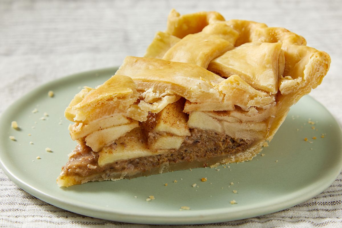 A slice of apple pie with a golden lattice crust sits on a light green plate, showing layers of sliced apples and cinnamon filling.