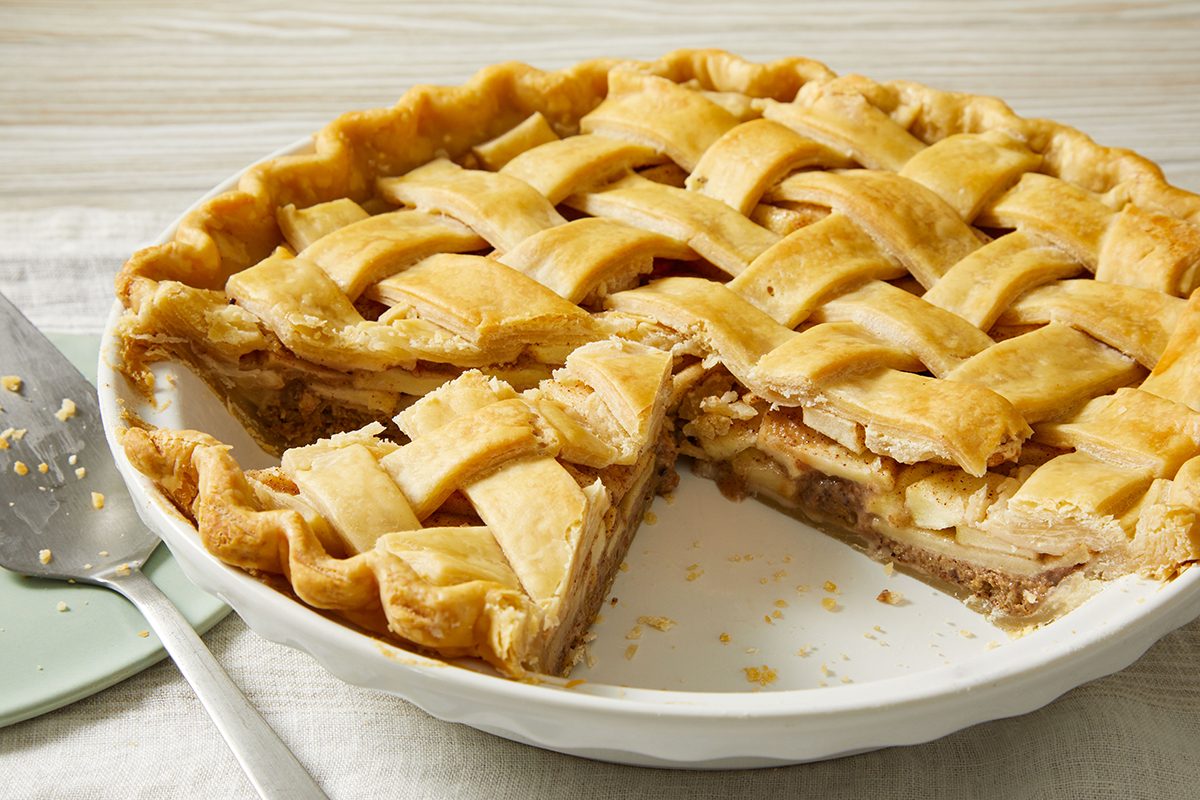 A lattice-topped apple pie with a slice removed, showing the filling, sits in a white pie dish on a light surface, with a pie server nearby.
