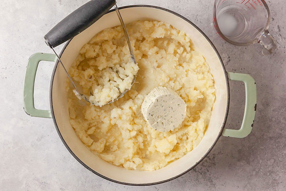 Overhead shot of a large pot filled with creamy mashed potatoes, topped with a round of herbed butter and a potato masher resting inside; A glass measuring cup with liquid sits beside the pot on a light surface;