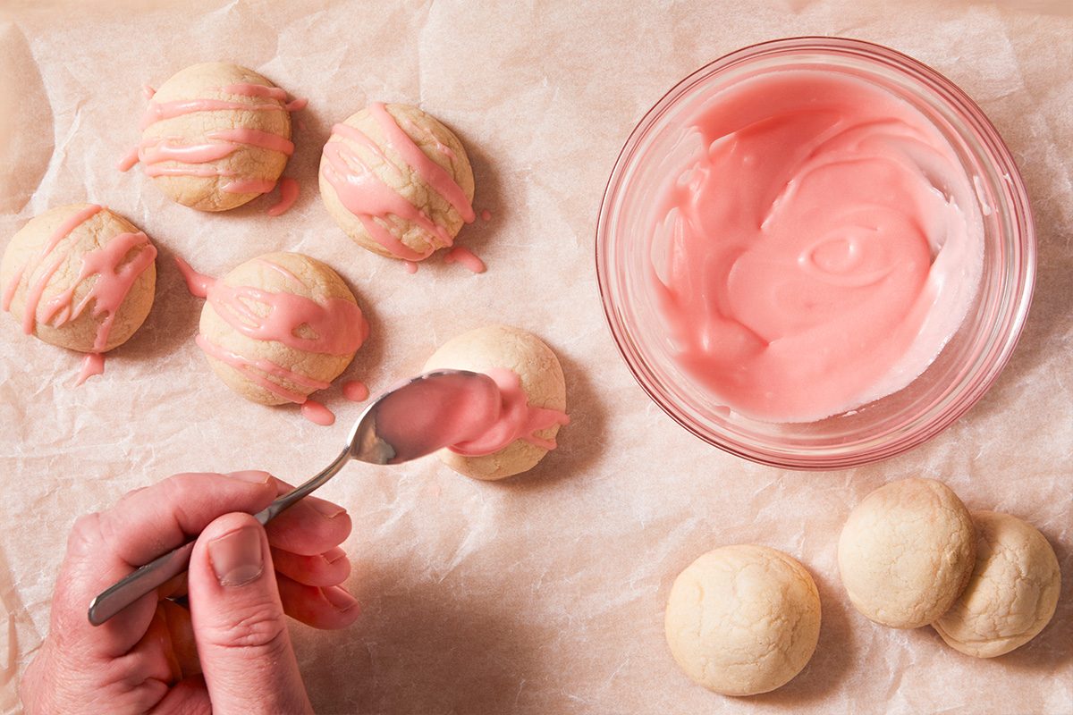 Overhead shot of baked Cherry Bonbon Cookies being drizzled with pink icing from a spoon beside a bowl of glaze.
