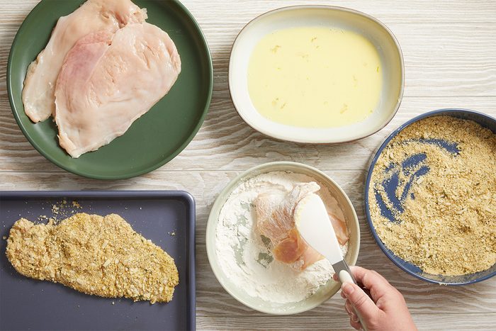 Overhead shot showing steps for breading chicken, flattened chicken breast dipped in egg wash and coated with breadcrumbs on plates.