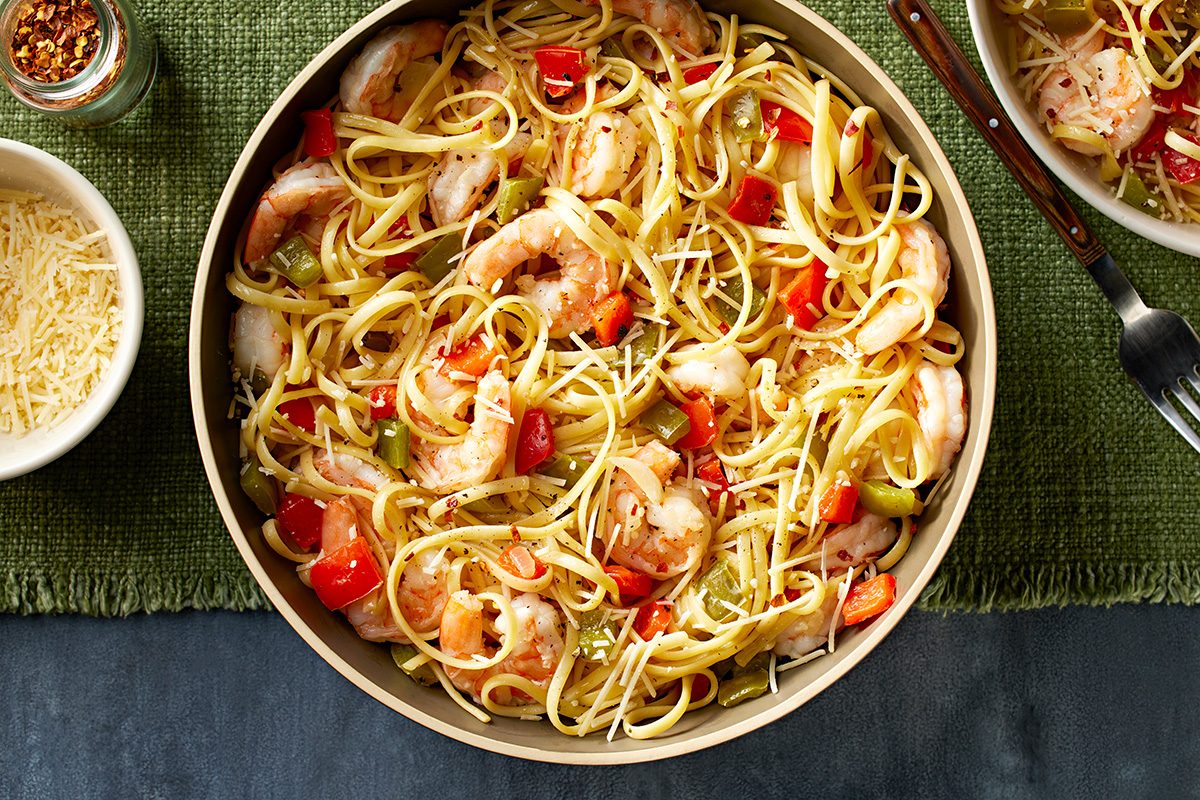 A bowl of shrimp pasta with linguine, cherry tomatoes, and herbs sits on a green placemat, with a small bowl of grated cheese and crushed red pepper nearby. A partial plate of pasta and a fork are also visible.