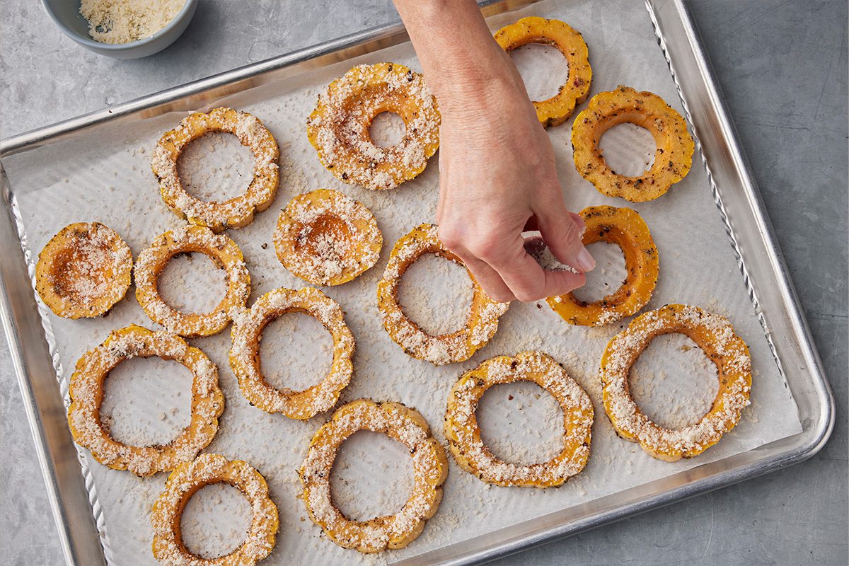 Overhead shot of a hand sprinkling breadcrumbs over sliced rings of squash arranged on a parchment-lined baking sheet;