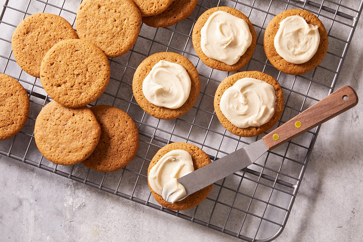Overhead shot of freshly baked ginger cookies on a wire rack, half spread with creamy frosting and a knife resting beside.