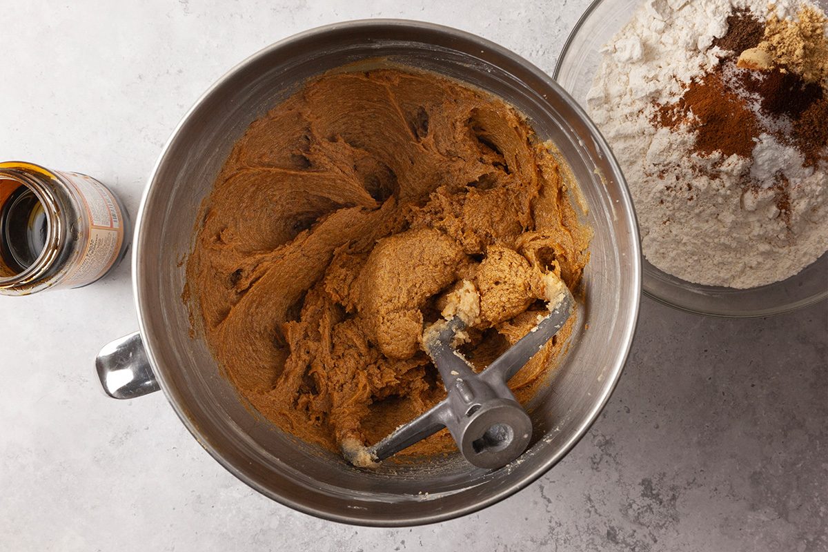 Overhead shot of a metal mixing bowl filled with brown cookie dough being mixed on a light countertop. Beside it sits a glass bowl containing flour and spices, and a small bottle of vanilla extract rests nearby;