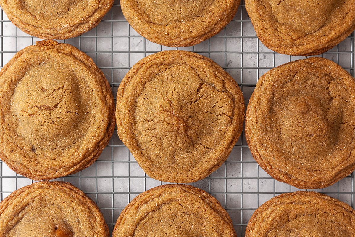 Overhead shot of Gingerbread Cheesecake Cookies cooling on a wire rack, each cookie golden brown with a domed center and slightly crinkled surface; The cookies are arranged in neat rows, showing their spiced, sugar-coated tops and soft, chewy texture;