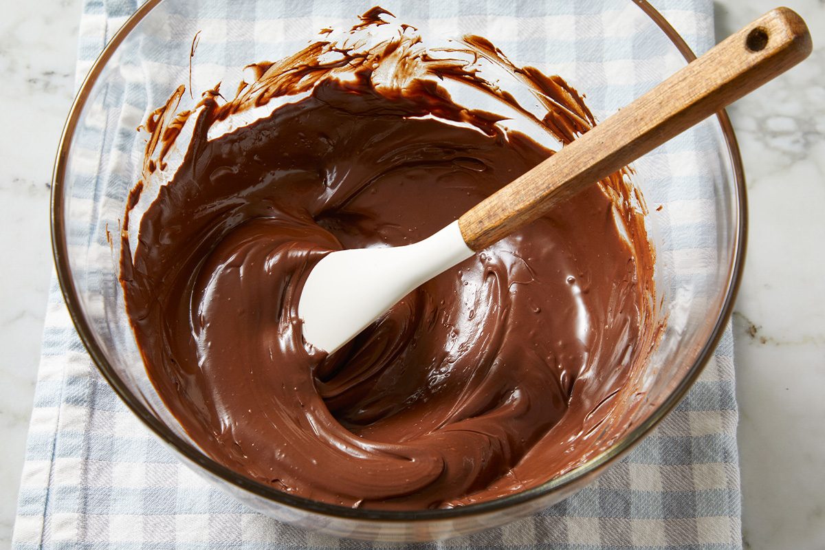 Overhead shot of melted chocolate being stirred with a spatula in a glass bowl on a blue checkered towel