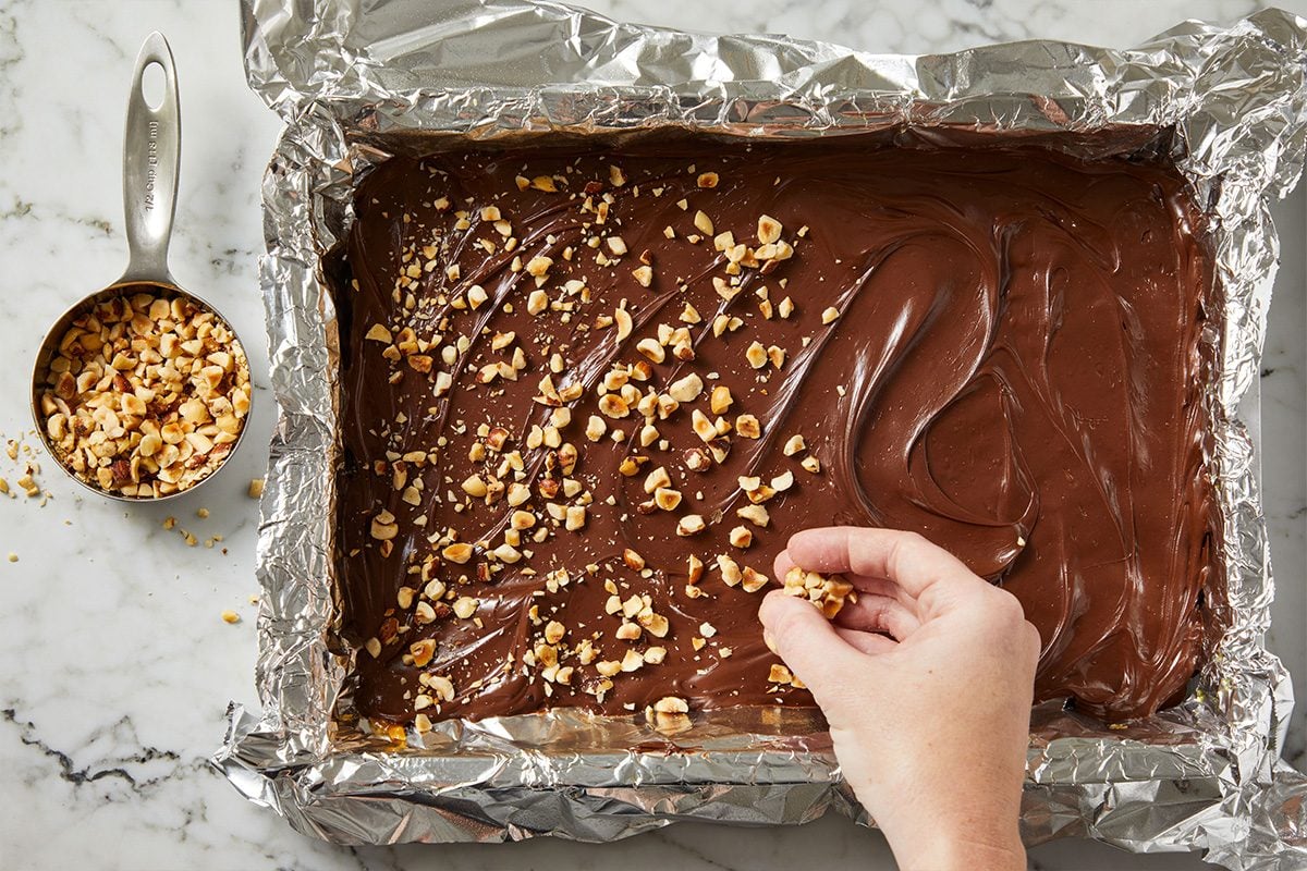 Overhead shot of chopped nuts being sprinkled over melted chocolate spread atop toffee in the pan.