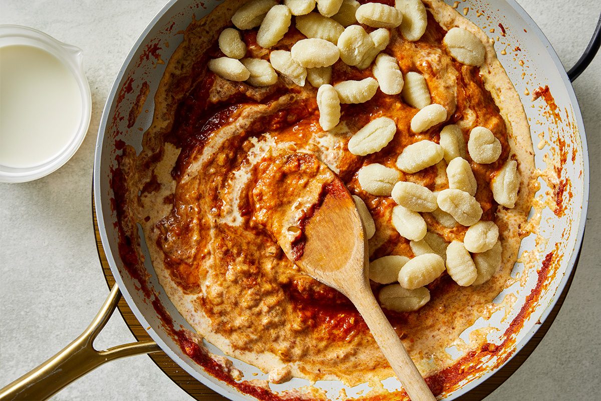 Overhead shot of a skillet filled with creamy tomato sauce being stirred with a wooden spoon, with uncooked gnocchi added on top; A small container of cream sits nearby on the counter;