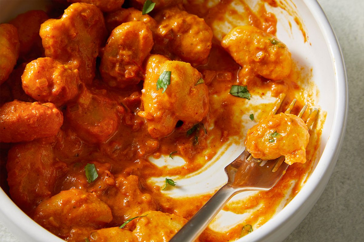 Closeup shot of a white bowl of Indian-Style Butter Gnocchi in a creamy orange sauce, topped with fresh herbs, with a fork lifting one gnocchi.