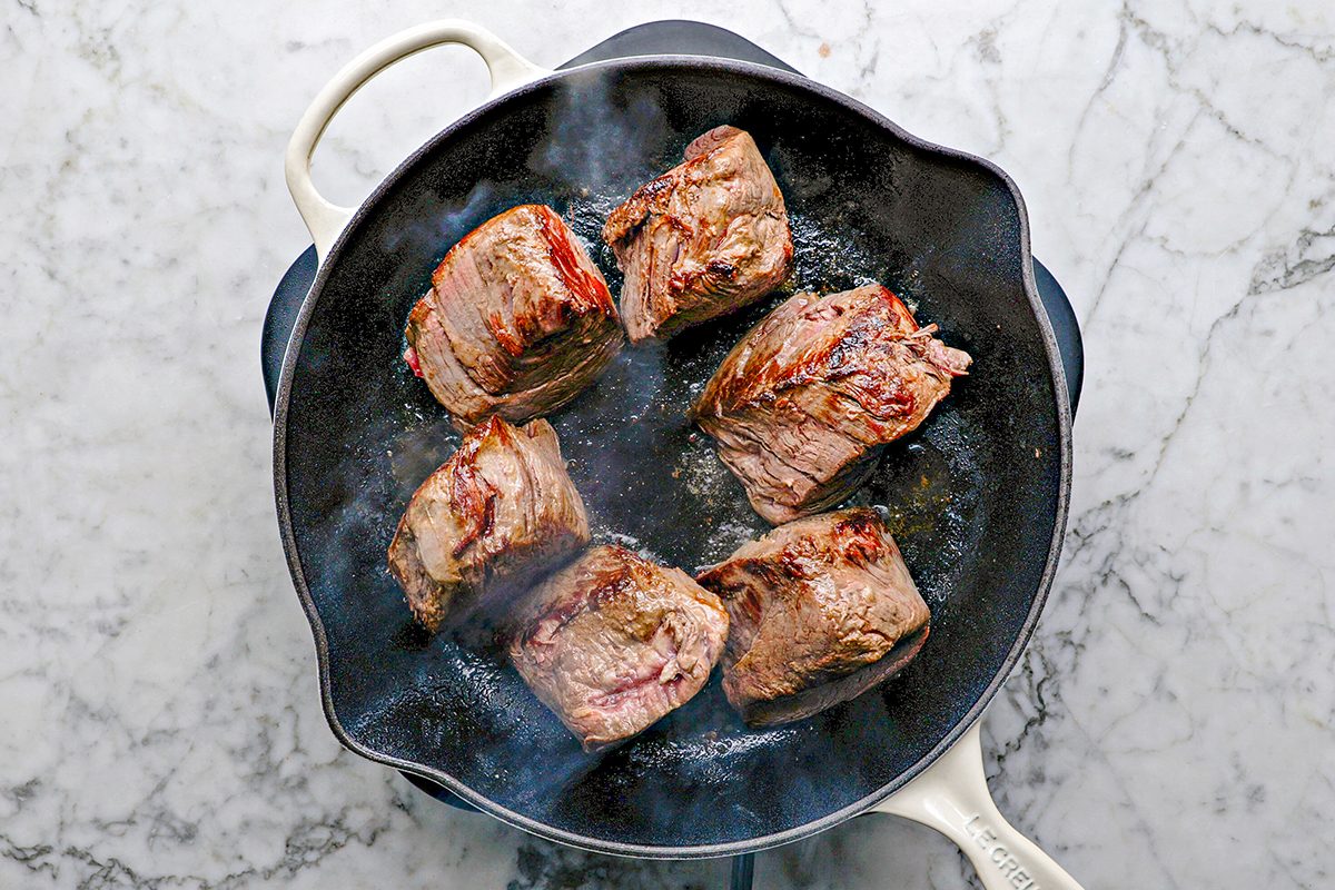 Overhead shot of six pieces of seared meat cooking in a black cast-iron skillet with a white handle, set on a white marble countertop; Steam rises from the pan;