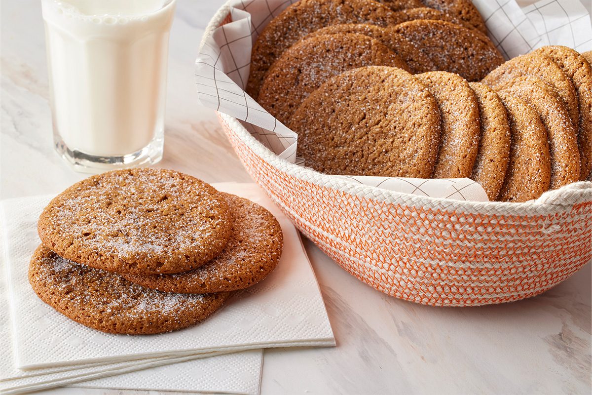 Table-view shot of a glass of milk beside two Joe Froggers on napkins, with a basket lined with patterned cloth holding more Joe Froggers in the background;