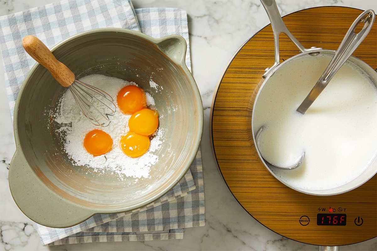 Overhead shot of a mixing bowl containing egg yolks and flour with a whisk, placed on a plaid cloth next to a saucepan of milk with a ladle, heated on an induction cooktop reading 178.6°F;