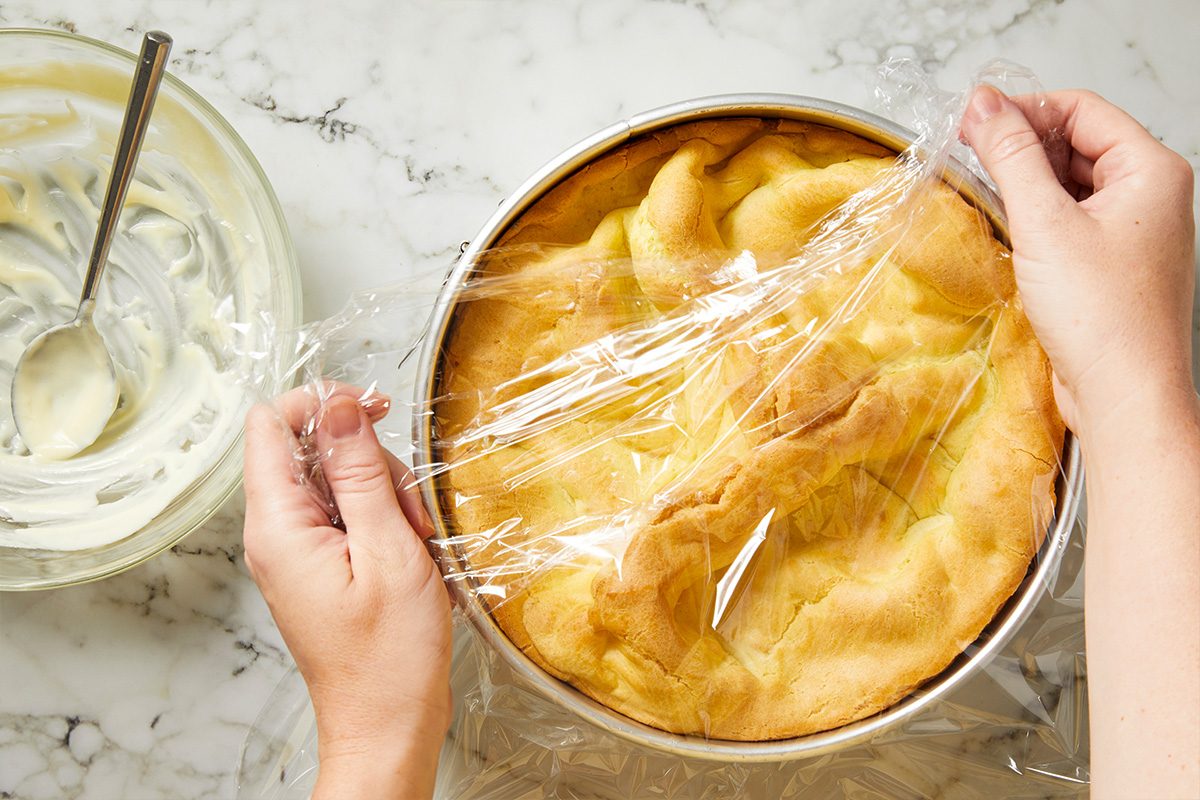 Overhead shot of hands covering a baked cake in a round pan with plastic wrap on a marble countertop, with a bowl containing a spoon and some creamy mixture nearby;