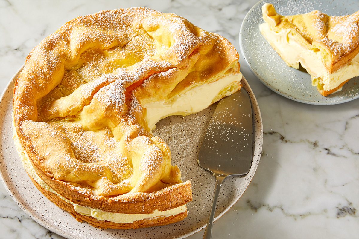 Overhead shot of Karpatka Cake with a slice removed, revealing thick creamy filling inside; The cake is dusted with powdered sugar and sits on a plate with a serving spatula;