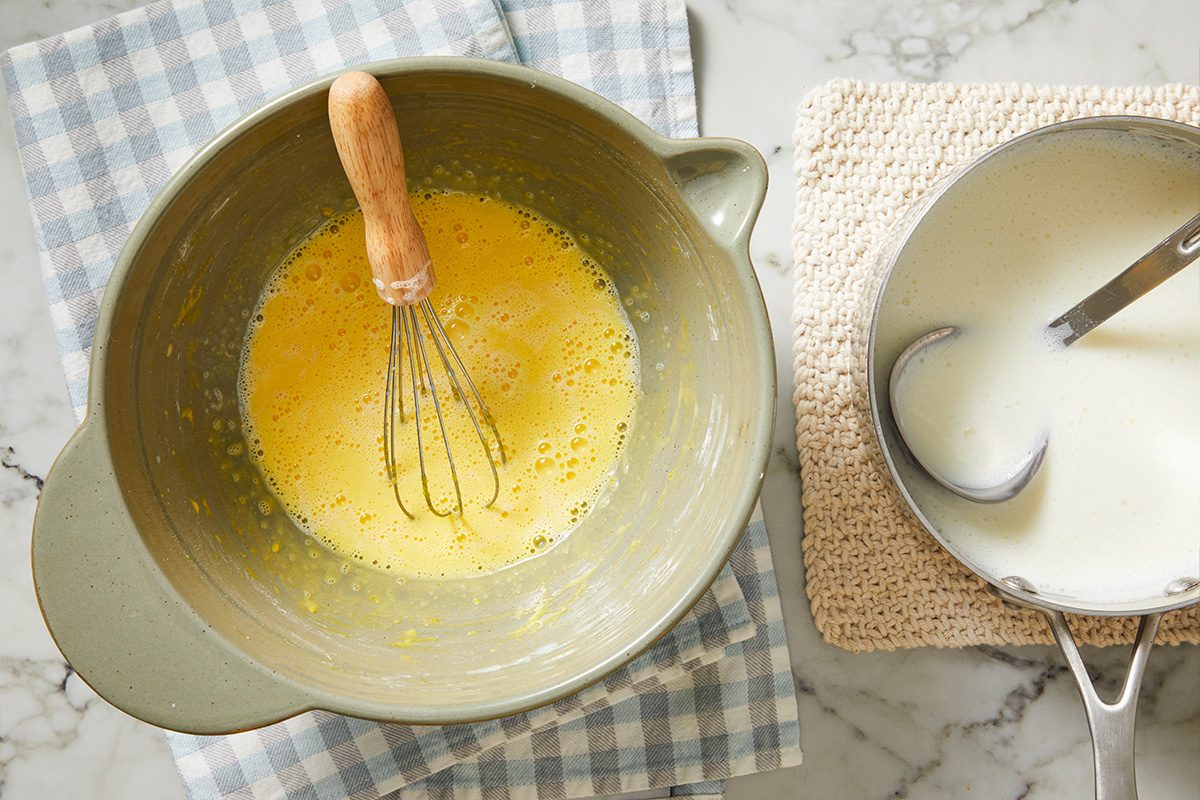 Overhead shot of a mixing bowl with a whisk and beaten eggs on a checkered cloth, placed next to a saucepan of creamy liquid with a ladle, both on a marble countertop;