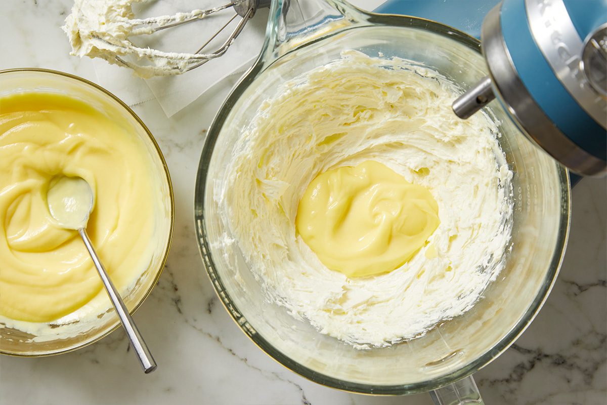 Overhead shot of two bowls on a marble countertop; one containing whipped cream and custard being mixed with an electric mixer, and the other holding yellow custard with a spoon; The mixer beaters rest nearby;
