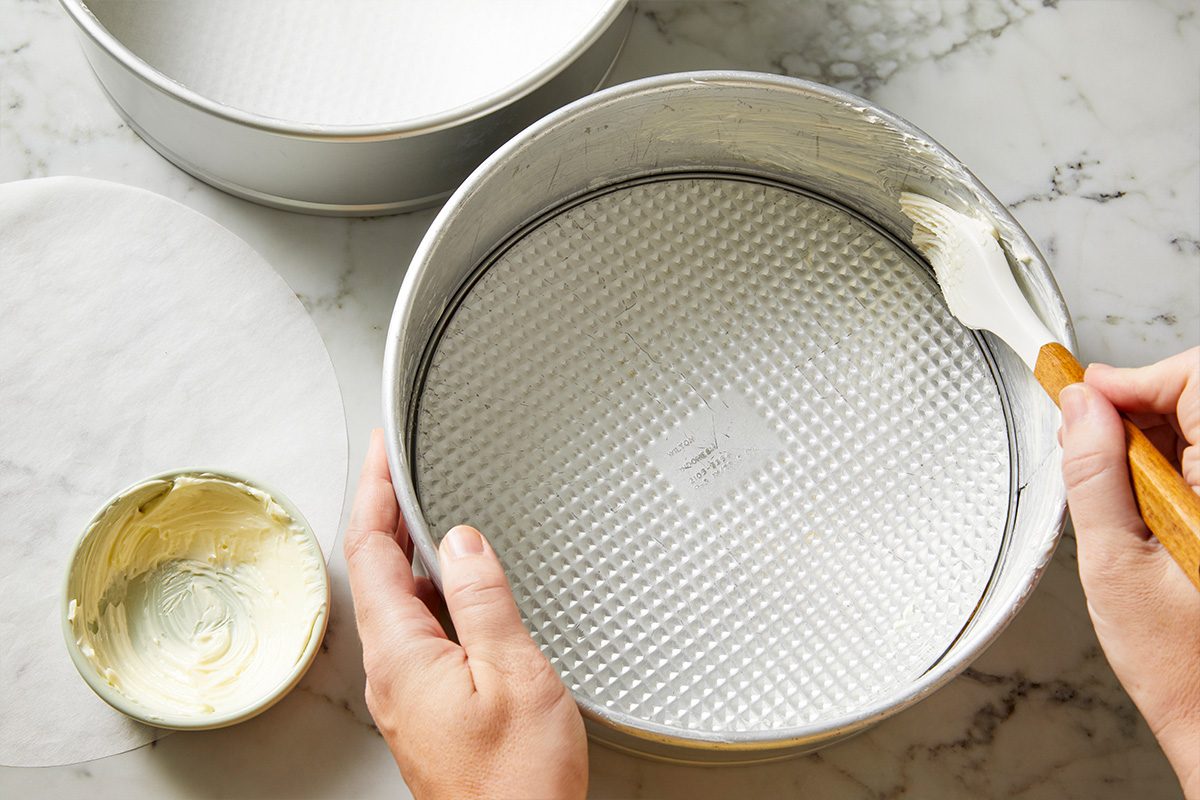 Overhead shot of a person using a brush to spread butter inside a round springform cake pan, with a small bowl of butter and a round piece of parchment paper nearby on a marble countertop;