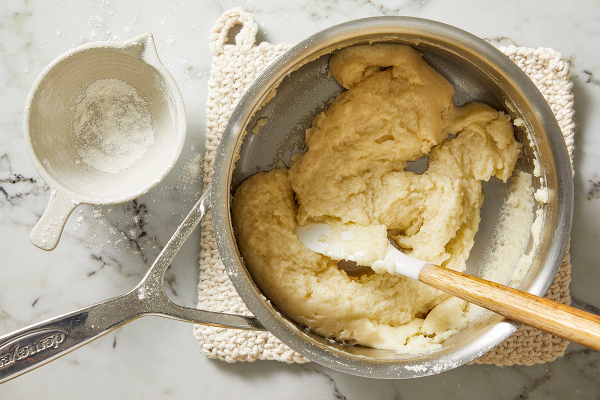 Overhead shot of a saucepan with dough being mixed by a spatula on a white knitted mat, with a small bowl of flour nearby, all on a marble countertop;