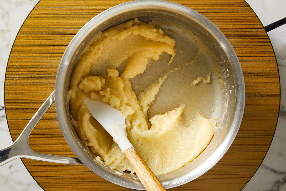 Overhead shot of a saucepan containing thick, pale dough being mixed with a white spatula with a wooden handle, placed on a round wooden surface;