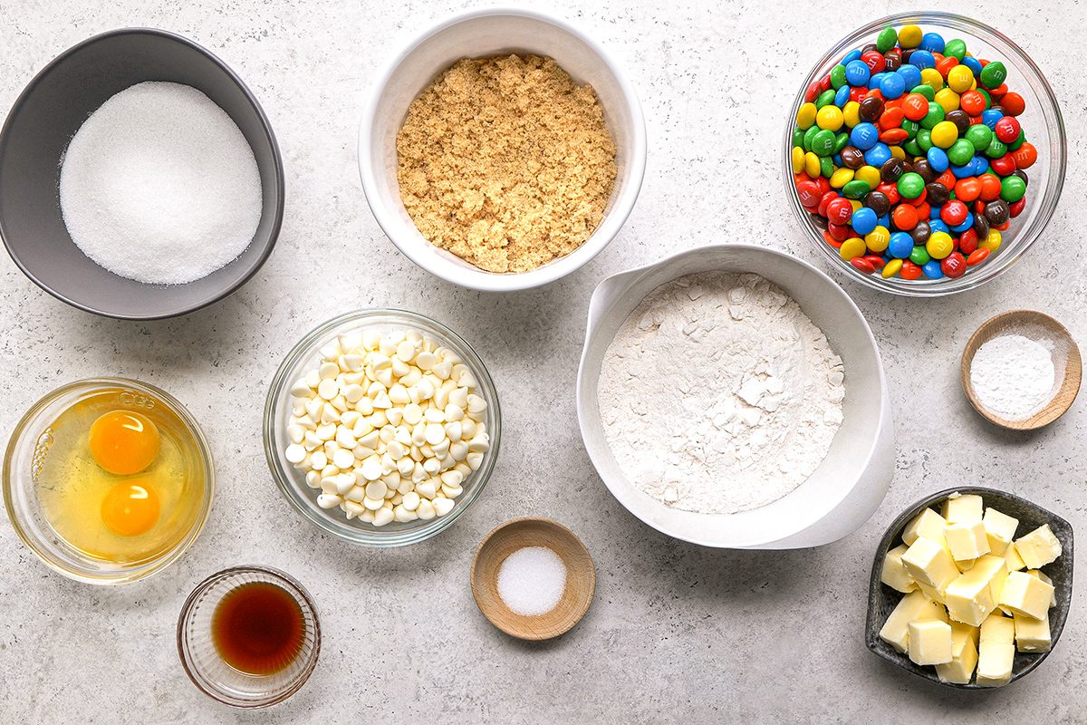 A top-down view of baking ingredients in bowls: sugar, crushed graham crackers, colorful candy-coated chocolates, eggs, white chocolate chips, flour, butter, vanilla extract, salt, and baking powder on a light surface.