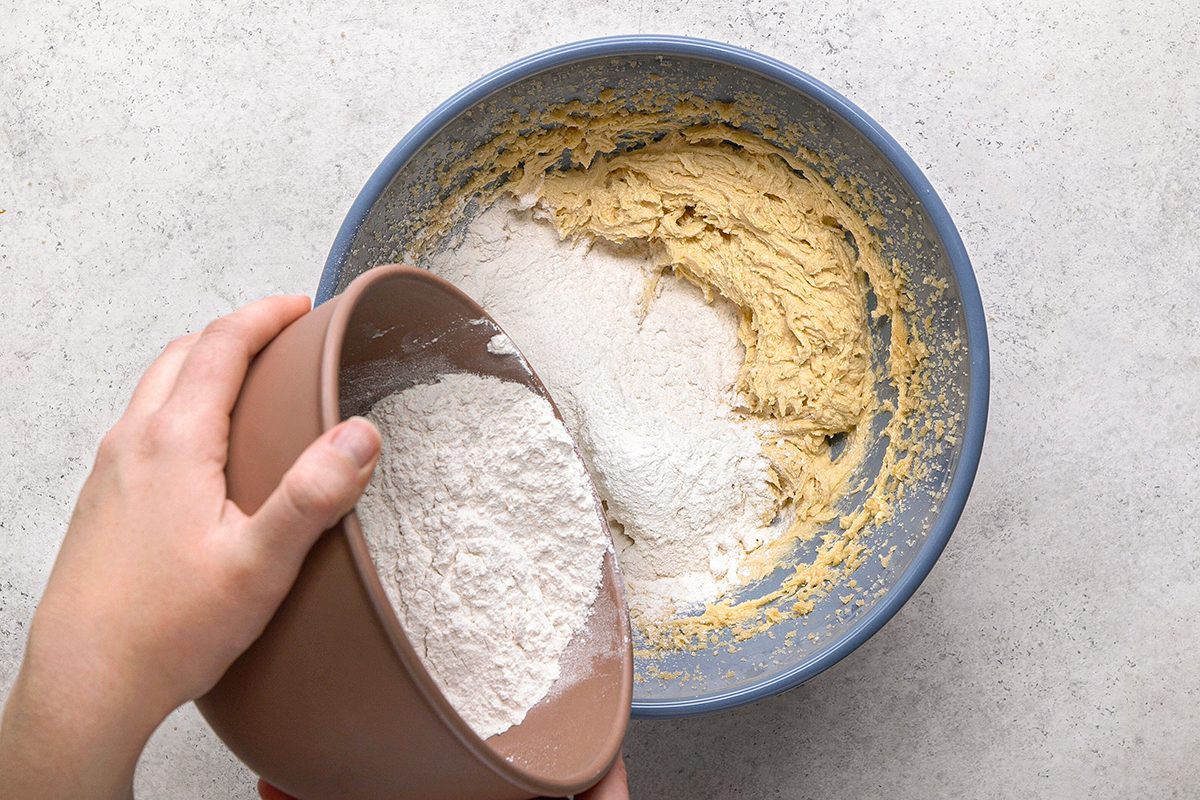A person pours flour from a brown bowl into a blue bowl containing partially mixed cookie dough on a light countertop.