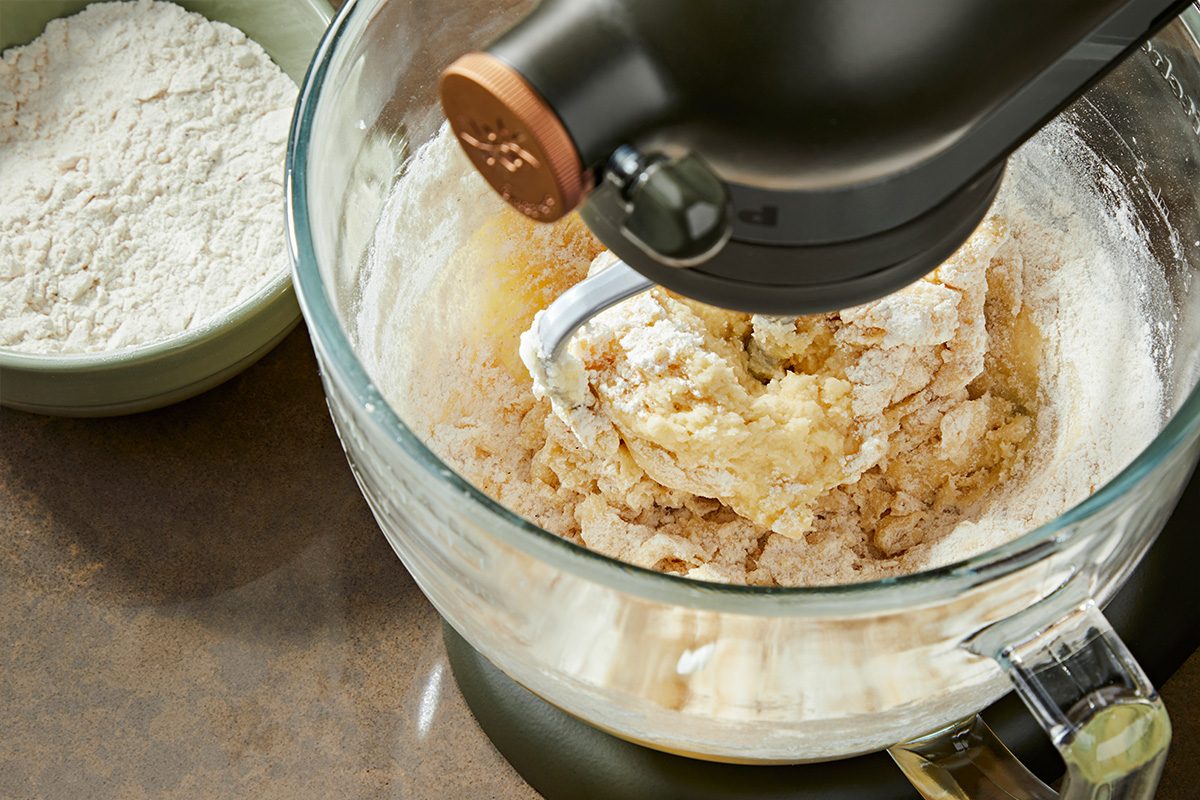 Closeup shot of a stand mixer with a glass bowl containing partially mixed dough and flour, with a green bowl of flour beside it on a brown countertop;