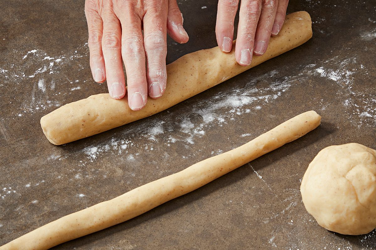 High angle view shot of hands rolling a piece of dough into a log on a floured surface, with another rolled dough log and a dough ball nearby;
