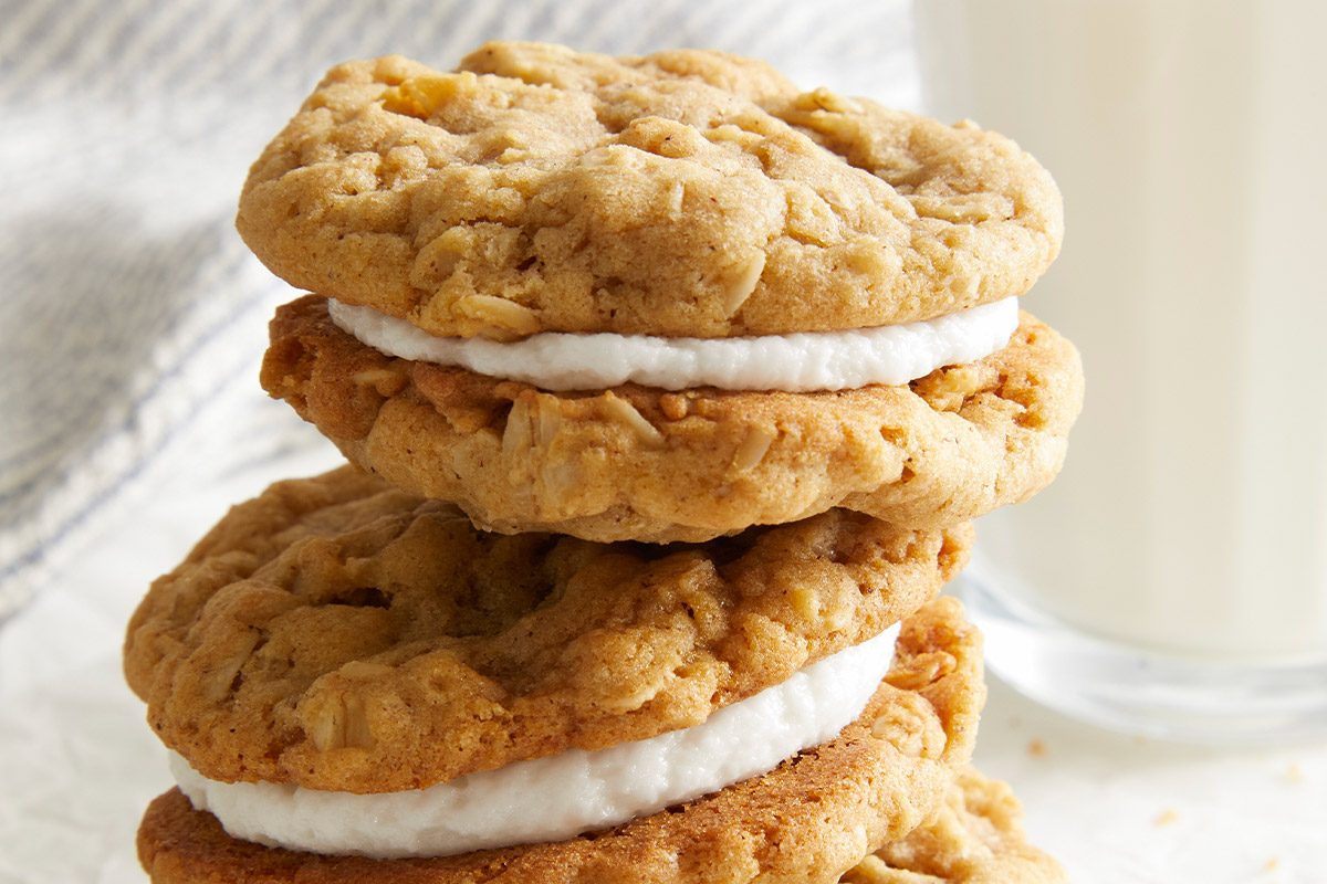 Vertical shot of Stacked Oatmeal Sandwich Cookies on a white surface, shown beside a glass of milk. Cream filling is clearly visible between the cookies.