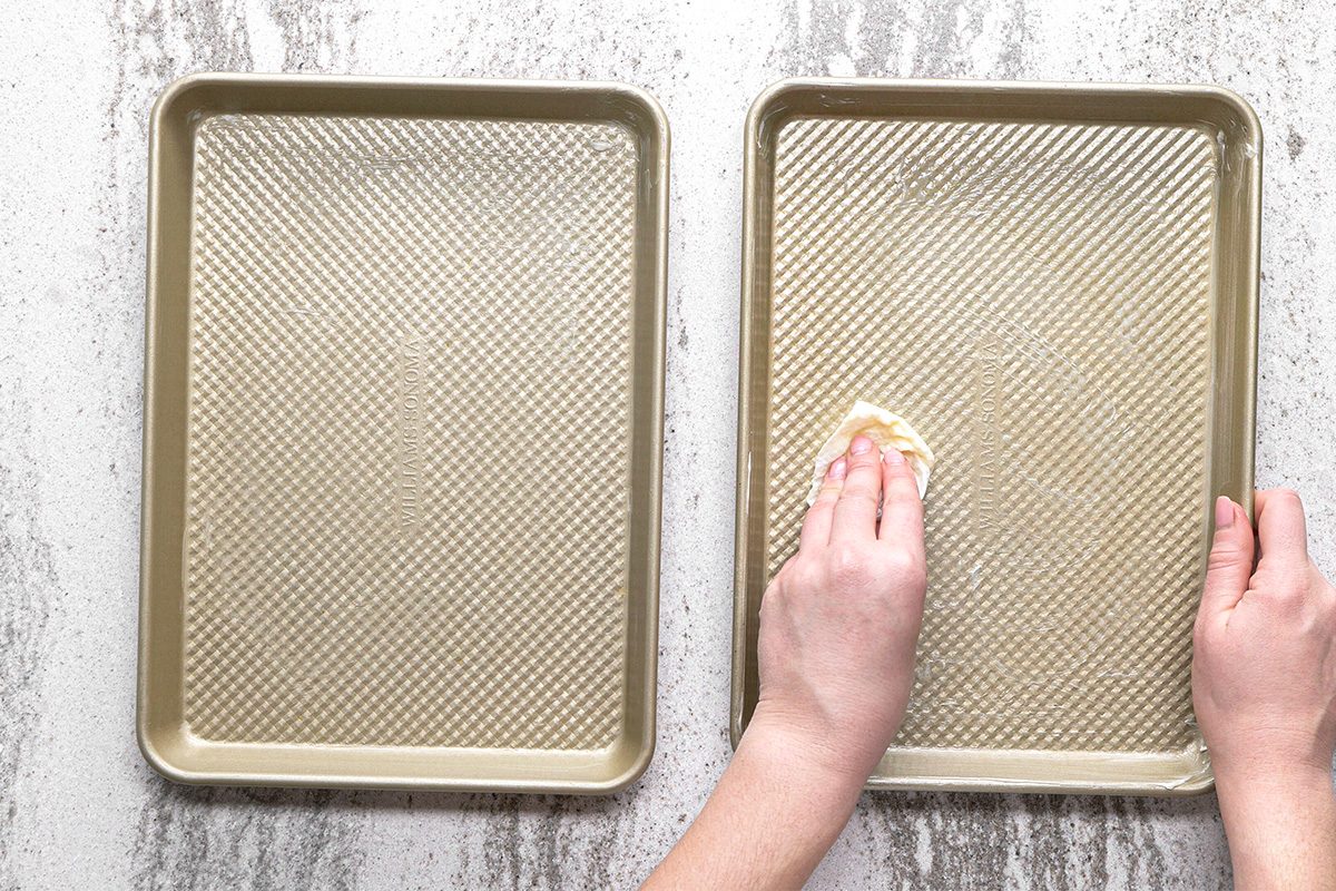 Two baking sheets are on a countertop. A hand is using a paper towel to grease one of the sheets, 
