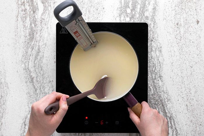 A person stirs a light-colored liquid in a saucepan on an induction cooktop, using a spatula. A candy thermometer is clipped to the pot, showing the temperature.