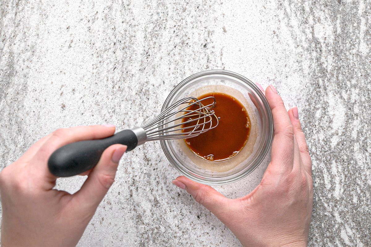 A person uses a whisk to mix a brown liquid in a small glass bowl on a light gray, speckled countertop.