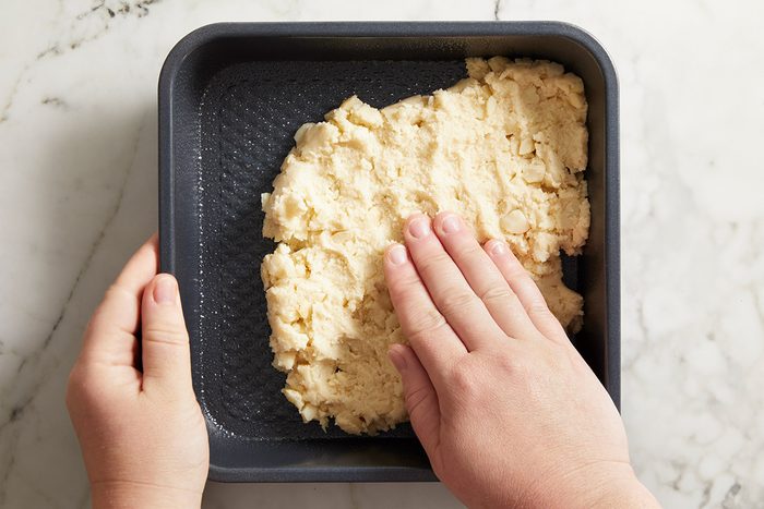 Overhead shot of hands pressing crust mixture evenly into a square baking pan lined with parchment paper.