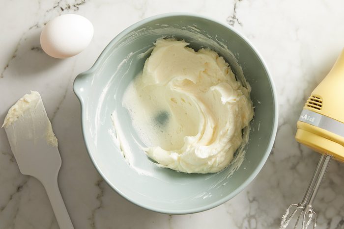 Overhead shot of cream cheese custard being mixed in a green bowl with an electric hand mixer, with an egg and spatula nearby.
