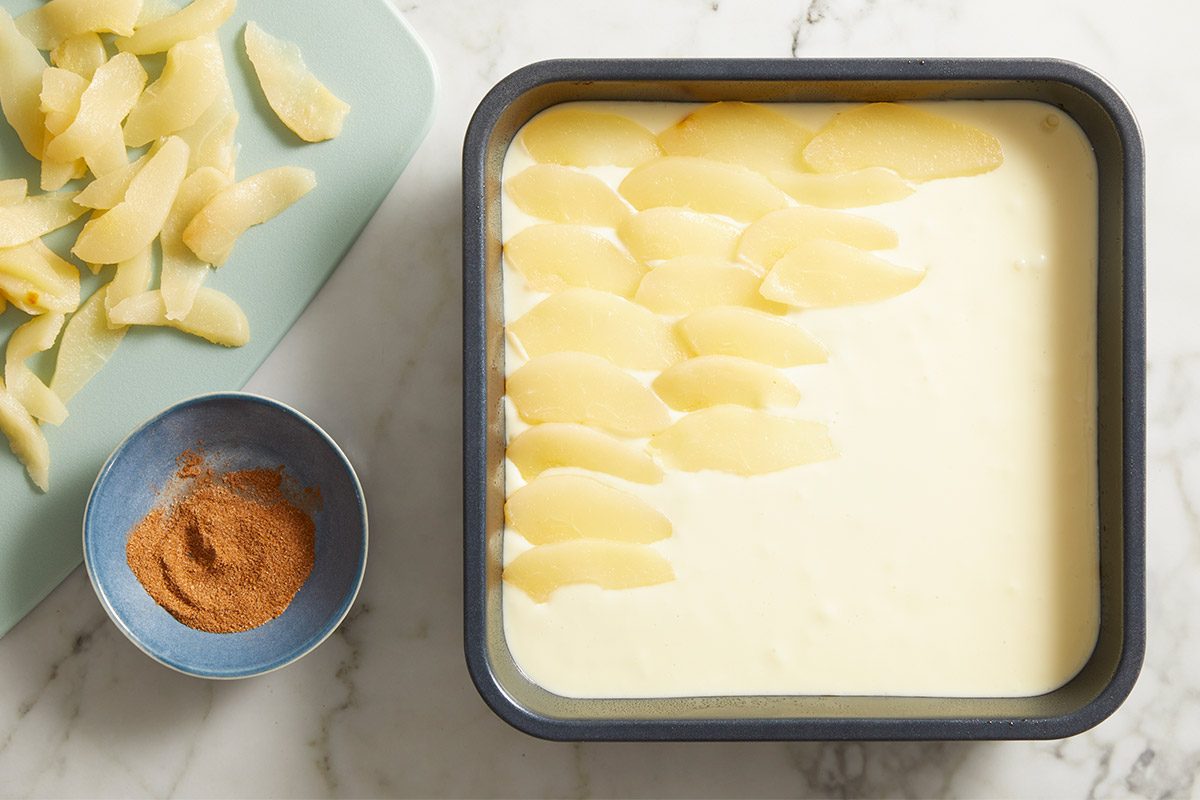 Overhead shot of thinly sliced pears arranged over custard filling in a square pan, with a small bowl of cinnamon sugar beside it.