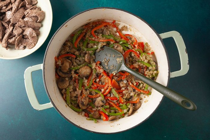 Overhead shot of a pot containing sautéed onions, mushrooms, and red and green bell peppers with a spatula resting inside, placed on a teal surface; Next to it sits a bowl filled with sliced cooked meat.