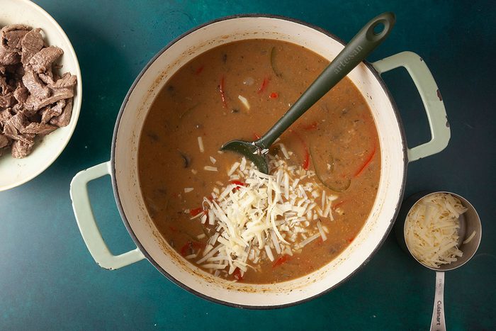 Overhead shot of a pot of soup topped with sliced vegetables and shredded cheese, with a ladle inside, sitting on a green surface; Nearby are a bowl of sliced cooked meat and a measuring cup filled with grated cheese;