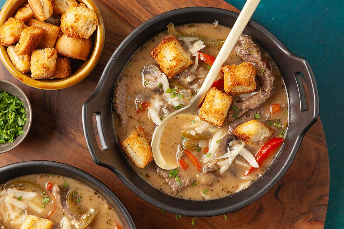 Overhead shot of two bowls of Philly Cheesesteak Soup filled with vegetables, meat, cheese, and croutons, set on a board with extra croutons, fresh herbs, and gold spoons nearby;