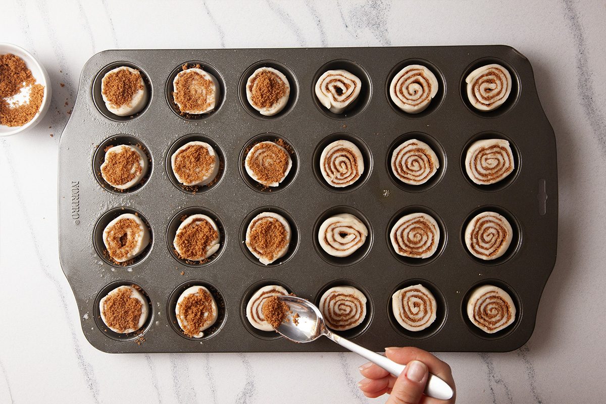 Overhead shot of a hand sprinkling a brown sugar and cinnamon mixture over unbaked mini cinnamon rolls arranged in a nonstick muffin tin, set on a white marble countertop;