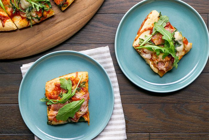 overhead shot of Two blue plates each hold a slice of pizza topped with arugula and prosciutto on a wooden table; A larger wooden board with more pizza is partially visible in the background; One plate sits on a striped napkin
