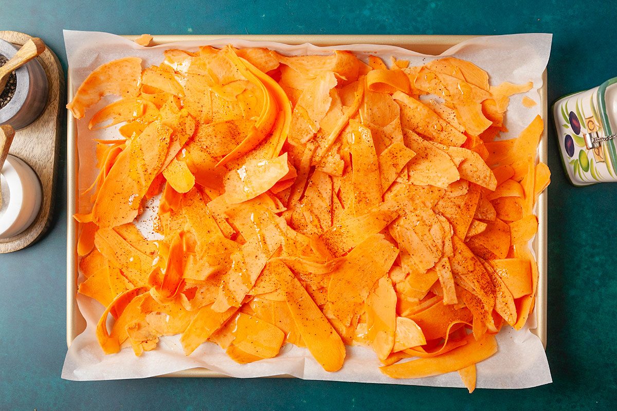 Overhead shot of thinly sliced sweet potato ribbons arranged on a parchment-lined baking tray, lightly seasoned with spices and ready to be roasted;