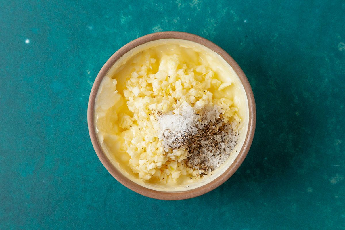 Overhead shot of a bowl filled with mashed potatoes, topped with minced garlic, grated cheese, salt, and pepper, placed on a teal surface;