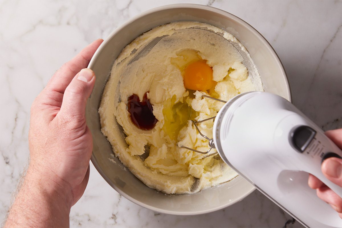 Overhead shot of a mixing bowl with butter, sugar, egg, and vanilla being blended using a hand mixer on a marble countertop.