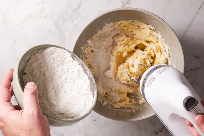Overhead view of creamed butter and sugar mixture being blended further with a hand mixer as flour is added from a small metal bowl.