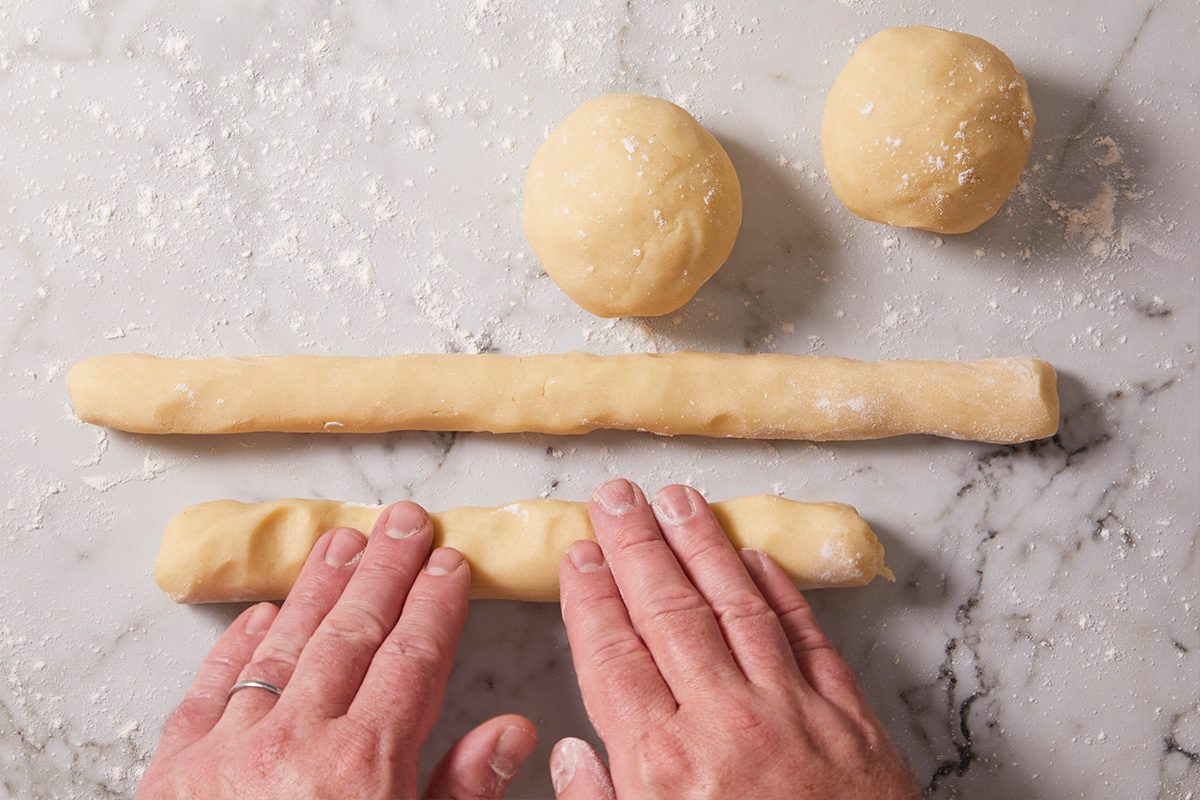 Overhead photo of hands shaping cookie dough into long logs on a floured surface with two dough balls visible above.