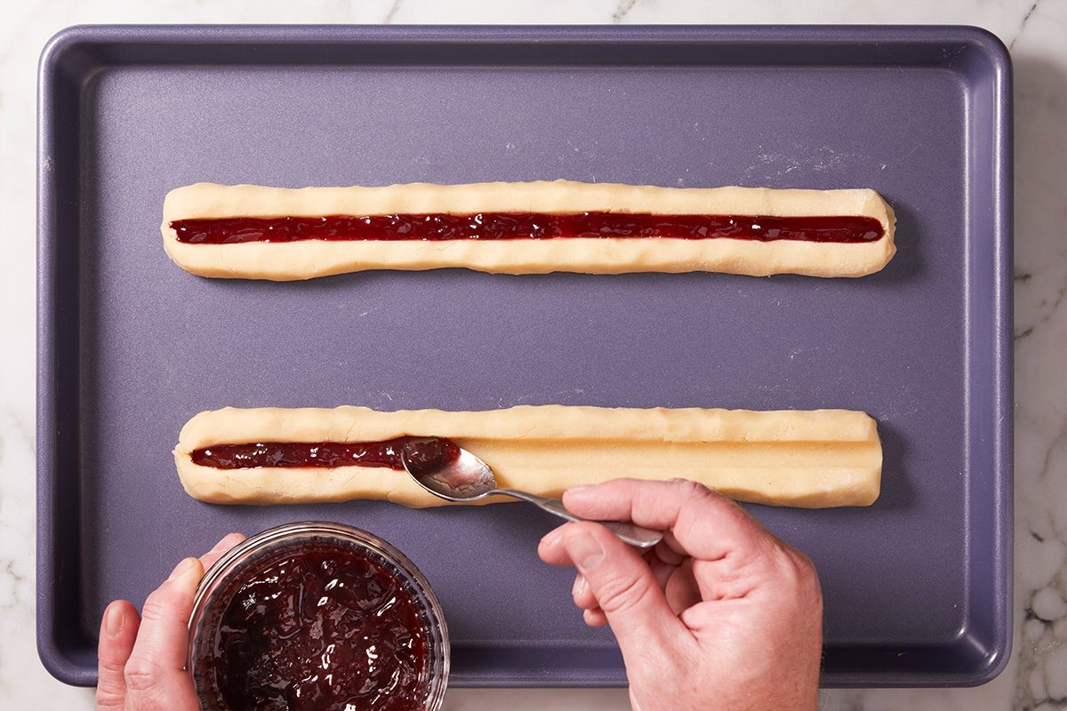 Split Second COverhead view of cookie logs on a baking sheet, with jam being spooned into the center grooves of the dough.ookies Ft25 6786 Jr 1008 4