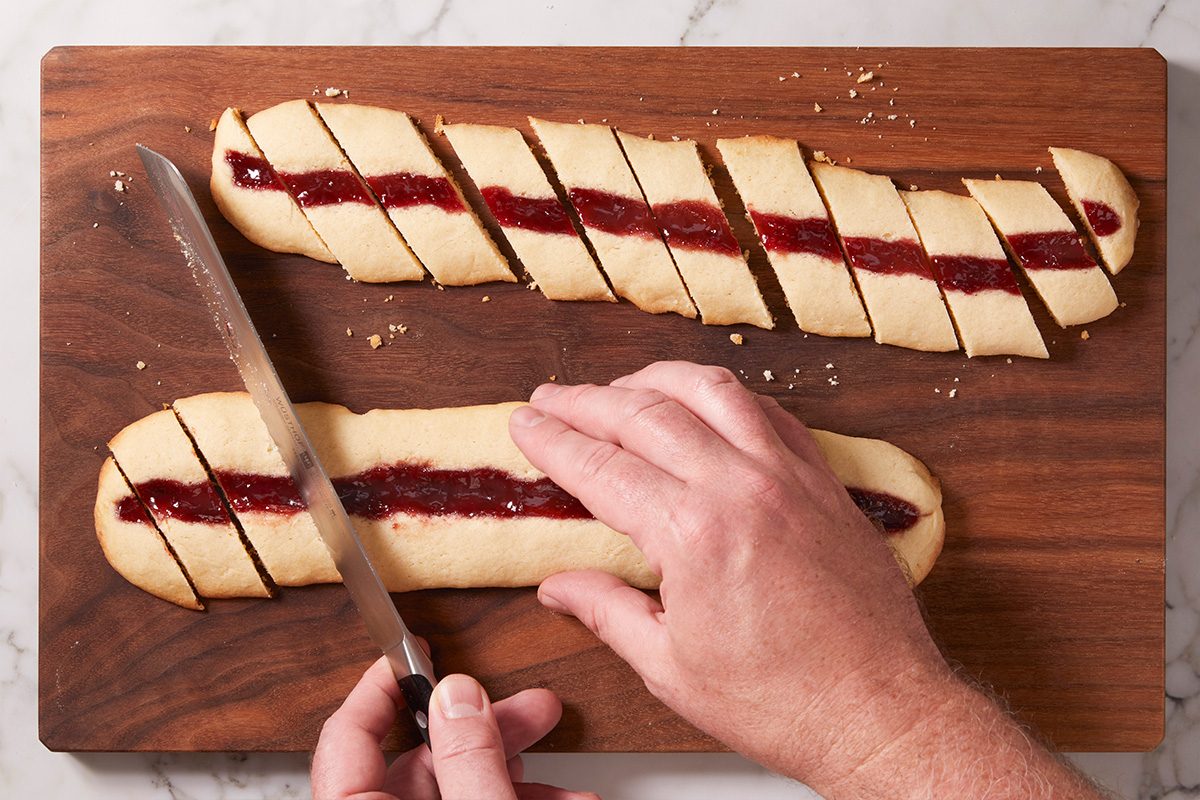 Overhead shot of hands slicing baked jam-filled cookie logs into diagonal pieces on a wooden cutting board.