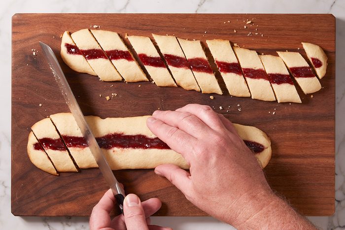 Overhead shot of hands slicing baked jam-filled cookie logs into diagonal pieces on a wooden cutting board.